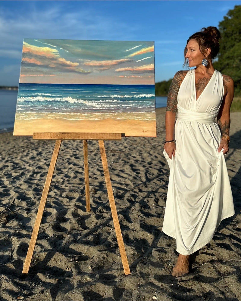 Woman in a white dress standing next to a beach scene painting on an easel by the water.